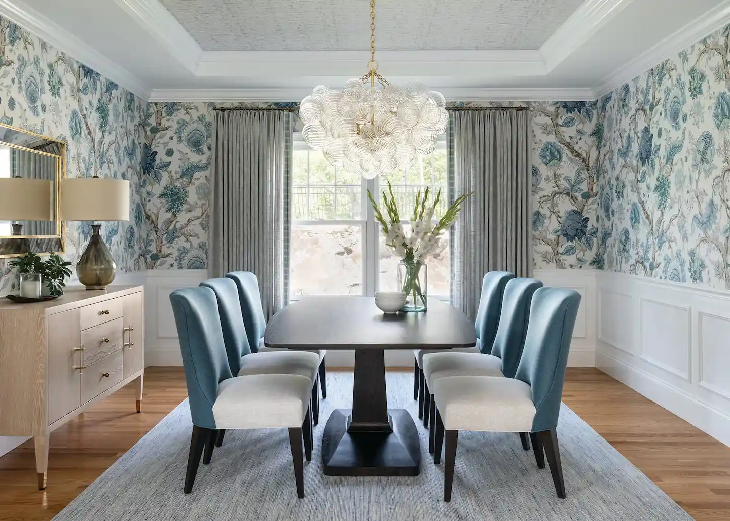 Formal dining room with pale blue botanical wallpaper, blue upholstered chairs, white wainscoting, and a sculptural pendant light.